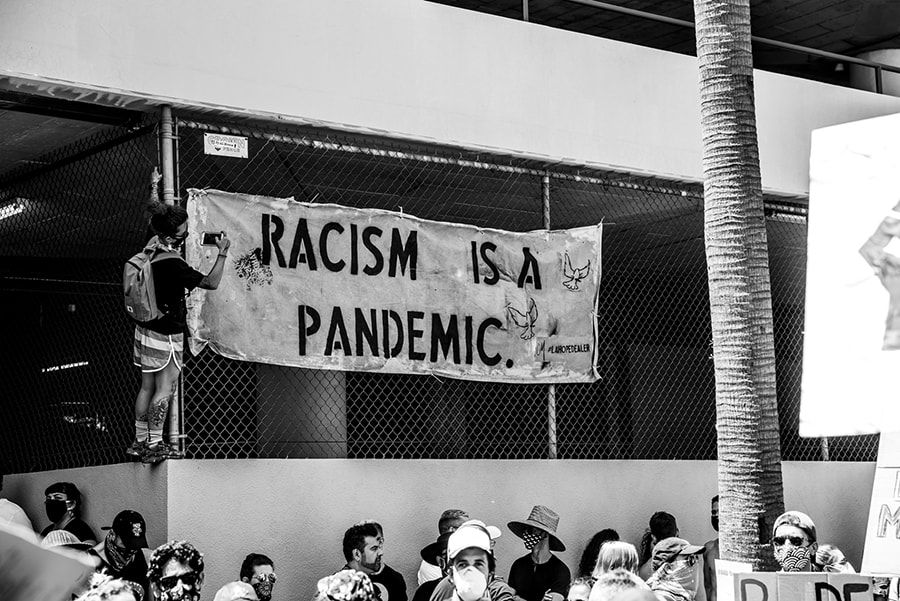 photograph of student sit in with students wearing masks. Banner reads "Racism is a Pandemic"